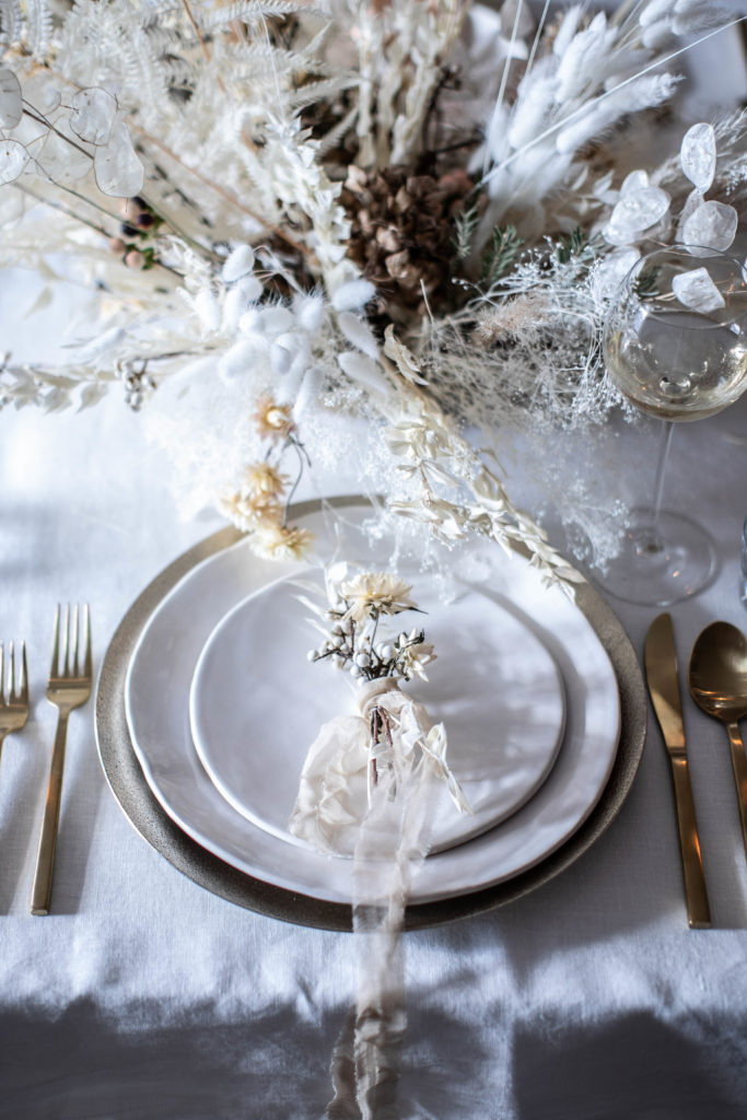 Minimalist, modern, neutral holiday tablescape for Christmas. A winter table setting with a natural, unique dried flower centerpiece, white linen table cloth, mini bouquet place settings, and brass gold flatware. The star is a dried flower bouquet and homemade wreaths with pampas grass, bleached fern, tallow berry, bunny tail grass, and lunaria. By Beth Kirby | Local Milk Blog.